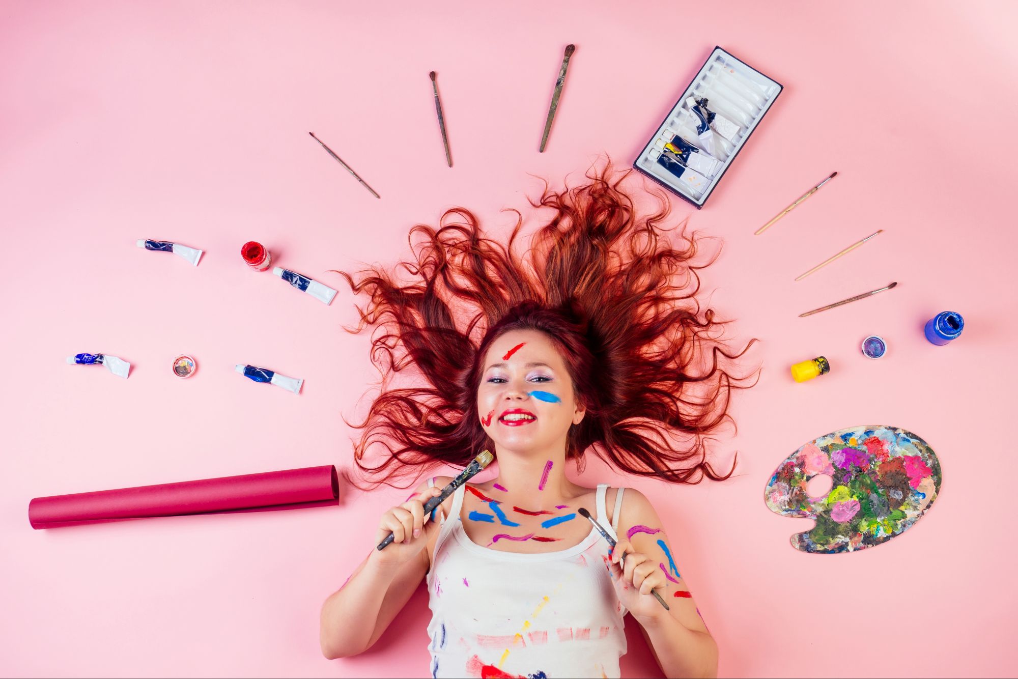 Joyful creative woman lying on pink background surrounded by paints and brushes, expressing creativity from God through color and bold artistic freedom.