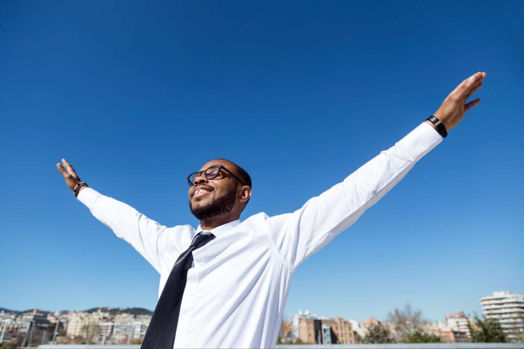 Man smiling with arms outstretched under clear blue sky, symbolizing bold faith and freedom in God