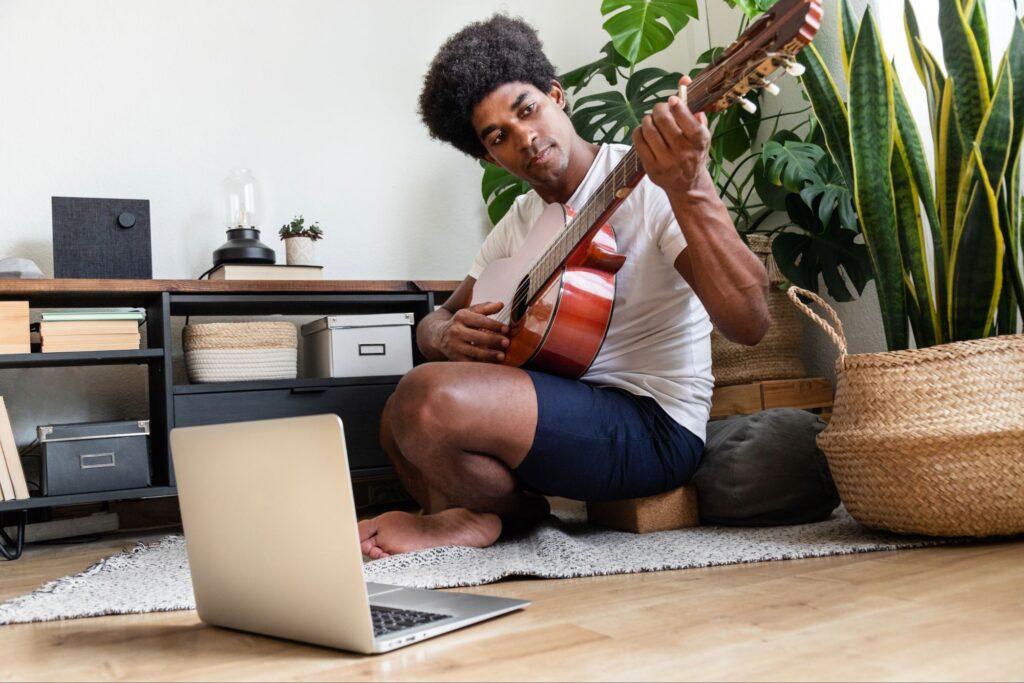 Focused young man playing guitar at home with laptop nearby, showing creativity from God through music and peaceful inspiration.